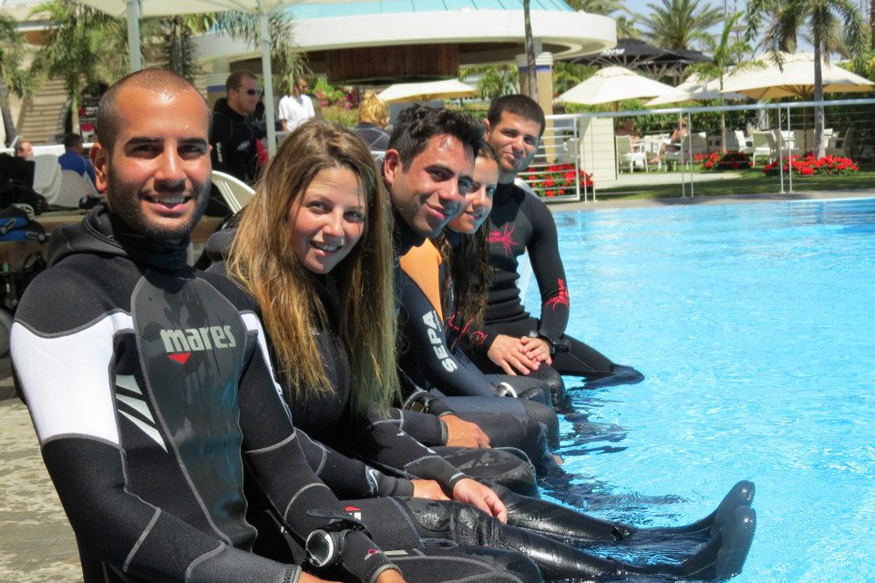 שורת צוללים בחליפות צלילה יושבים בתור על שפת הבריכה עם רגליים במים